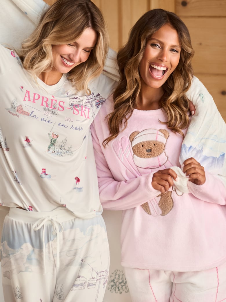 Two models by a fireplace, one in pink teddy pajamas, the other in white ski-patterned pajamas.