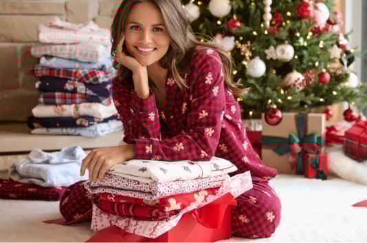 Mannequin assise devant un sapin de Noël en pyjama rouge à carreaux, préparant une pile de pyjamas à emballer.